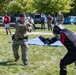 A military training exercise is taking place outdoors on a grassy area. A sailor in Navy Working Uniform (NWU) camouflage is pointing a training firearm at a person wearing full protective padded training gear (red and black suit with helmet). Several observers, including military personnel and civilians, watch from the background.
