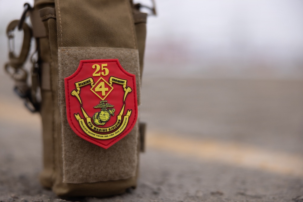 A close-up photograph of a PVC military patch affixed to a tan/coyote brown MOLLE tactical pouch. The patch belongs to the 25th Marine Regiment, 4th Marine Division, featuring the Eagle, Globe, and Anchor emblem. The background is blurred, suggesting an outdoor environment with a road or tarmac surface visible.