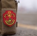 A close-up photograph of a PVC military patch affixed to a tan/coyote brown MOLLE tactical pouch. The patch belongs to the 25th Marine Regiment, 4th Marine Division, featuring the Eagle, Globe, and Anchor emblem. The background is blurred, suggesting an outdoor environment with a road or tarmac surface visible.
