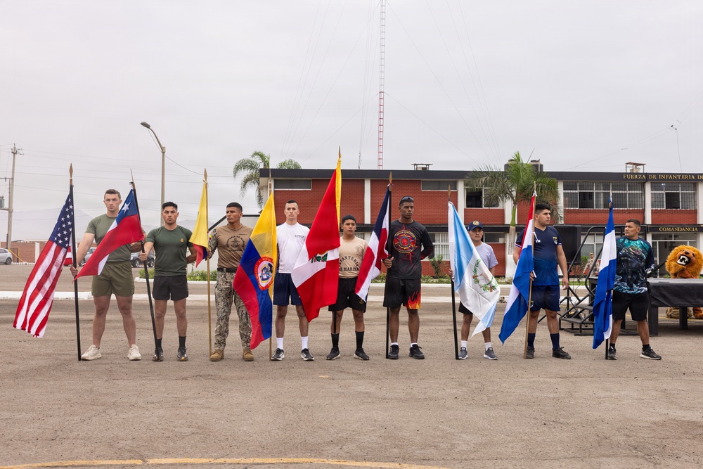 A group of approximately ten individuals stand in a line on a paved parade ground, each holding a national flag. The setting appears to be a military installation, as indicated by the building sign reading 'FUERZA DE INFANTERIA DE' and 'COMANDANCIA' in the background. This appears to be a multinational military ceremony or event involving personnel from multiple Latin American countries and the United States.