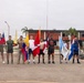 A group of approximately ten individuals stand in a line on a paved parade ground, each holding a national flag. The setting appears to be a military installation, as indicated by the building sign reading 'FUERZA DE INFANTERIA DE' and 'COMANDANCIA' in the background. This appears to be a multinational military ceremony or event involving personnel from multiple Latin American countries and the United States.