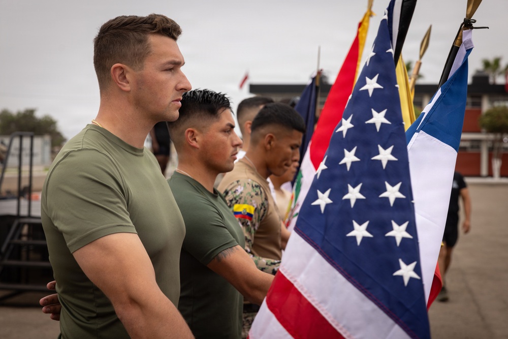 A group of military personnel in olive green t-shirts and camouflage uniforms stand at attention in a formal formation. Multiple national flags are visible, including the United States flag prominently in the foreground and what appears to be the Ecuadorian flag patch on one soldier's uniform. The setting appears to be an outdoor military installation or parade ground.