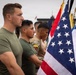 A group of military personnel in olive green t-shirts and camouflage uniforms stand at attention in a formal formation. Multiple national flags are visible, including the United States flag prominently in the foreground and what appears to be the Ecuadorian flag patch on one soldier's uniform. The setting appears to be an outdoor military installation or parade ground.