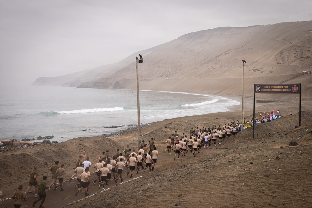 A large group of military personnel in uniform are running along a coastal dirt path toward a gate/arch structure. The sign on the arch reads 'Área de Entrenamiento para Operaciones de Búsqueda y Rescate' (Search and Rescue Operations Training Area), indicating this is a military training facility. The setting features a dramatic arid coastal landscape with steep sandy cliffs, ocean waves, and overcast skies consistent with the Peruvian coast.