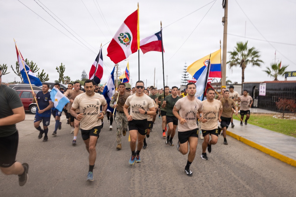 A large group of military personnel in matching tan shirts and black shorts are running together down a road while carrying multiple national flags. The shirts appear to read 'Infantería de Marina' (Marine Infantry), suggesting this is a multinational military exercise or ceremonial run. Multiple Latin American national flags are visible, including those of Peru, Chile, Colombia, Argentina, Dominican Republic, and Honduras.