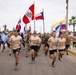 A large group of military personnel in matching tan shirts and black shorts are running together down a road while carrying multiple national flags. The shirts appear to read 'Infantería de Marina' (Marine Infantry), suggesting this is a multinational military exercise or ceremonial run. Multiple Latin American national flags are visible, including those of Peru, Chile, Colombia, Argentina, Dominican Republic, and Honduras.