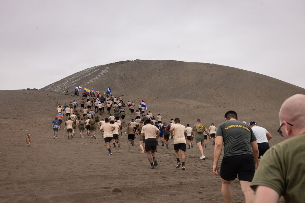 A large group of military personnel from multiple nations are running up a dark volcanic or sandy hill/dune. Several participants are carrying national flags, and one person in the foreground wears a shirt reading 'Infanteria de Marina' (Marine Infantry). The event appears to be a multinational military fitness or ceremonial run.