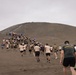 A large group of military personnel from multiple nations are running up a dark volcanic or sandy hill/dune. Several participants are carrying national flags, and one person in the foreground wears a shirt reading 'Infanteria de Marina' (Marine Infantry). The event appears to be a multinational military fitness or ceremonial run.