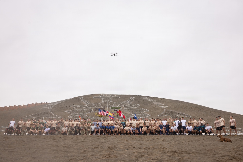 A large group of military personnel and civilians pose for a group photograph at the base of a sandy hill featuring a large geoglyph or emblem etched into its surface. Multiple national flags are visible among the group, suggesting a multinational military exercise or joint operation. A drone is visible in the overcast sky above, likely capturing aerial footage of the event.