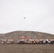 A large group of military personnel and civilians pose for a group photograph at the base of a sandy hill featuring a large geoglyph or emblem etched into its surface. Multiple national flags are visible among the group, suggesting a multinational military exercise or joint operation. A drone is visible in the overcast sky above, likely capturing aerial footage of the event.