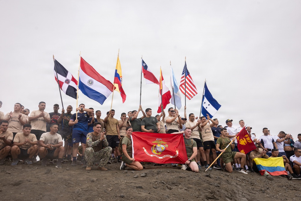A large group of military personnel from multiple nations pose together on elevated dirt terrain, holding their respective national flags. Two individuals in the foreground kneel while holding a red United States Marine Corps banner. The gathering appears to be a multinational military exercise or joint training event.