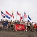 A large group of military personnel from multiple nations pose together on elevated dirt terrain, holding their respective national flags. Two individuals in the foreground kneel while holding a red United States Marine Corps banner. The gathering appears to be a multinational military exercise or joint training event.