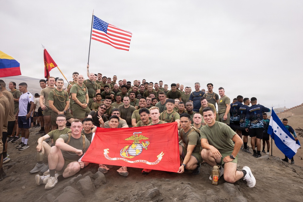 A large group of United States Marines and allied military personnel pose for a group photo on an elevated sandy terrain, likely during a multinational military exercise or training event. The group is holding a red United States Marine Corps flag in the foreground, along with an American flag, a Colombian flag, and a Honduran flag visible in the background. The participants are predominantly wearing olive drab military PT (physical training) shirts.