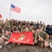 A large group of United States Marines and allied military personnel pose for a group photo on an elevated sandy terrain, likely during a multinational military exercise or training event. The group is holding a red United States Marine Corps flag in the foreground, along with an American flag, a Colombian flag, and a Honduran flag visible in the background. The participants are predominantly wearing olive drab military PT (physical training) shirts.