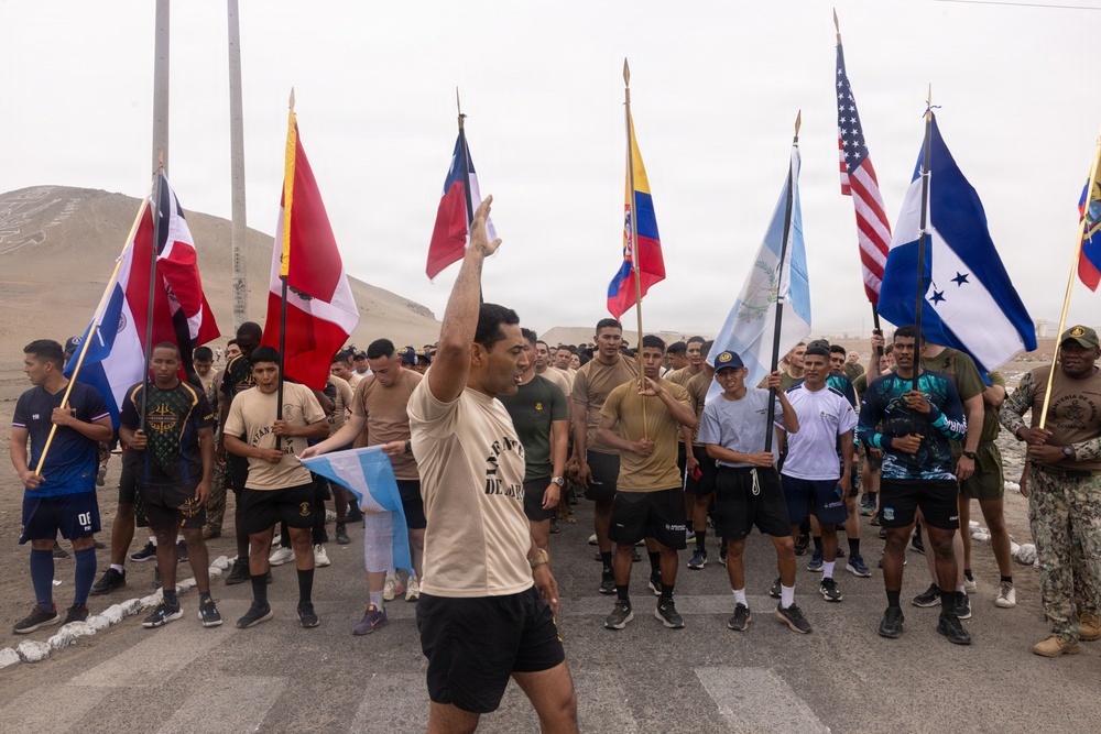 A large group of military personnel and participants are gathered outdoors in what appears to be a desert or arid environment, holding national flags from multiple countries. A central figure in a tan shirt is gesturing with his arm raised, appearing to lead or address the group. The event appears to be a multinational military exercise or ceremonial gathering.