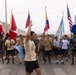 A large group of military personnel and participants are gathered outdoors in what appears to be a desert or arid environment, holding national flags from multiple countries. A central figure in a tan shirt is gesturing with his arm raised, appearing to lead or address the group. The event appears to be a multinational military exercise or ceremonial gathering.