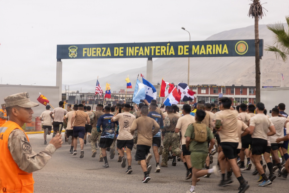 A large group of military personnel in various uniforms are running through the entrance gate of the 'Fuerza de Infantería de Marina' (Marine Infantry Force), a Peruvian naval installation. Participants are carrying multiple national flags including those of the United States, Honduras, Dominican Republic, Peru, Venezuela, and Ecuador, suggesting a multinational military exercise or ceremonial run. A safety marshal in an orange vest stands to the left observing the event.