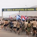 A large group of military personnel in various uniforms are running through the entrance gate of the 'Fuerza de Infantería de Marina' (Marine Infantry Force), a Peruvian naval installation. Participants are carrying multiple national flags including those of the United States, Honduras, Dominican Republic, Peru, Venezuela, and Ecuador, suggesting a multinational military exercise or ceremonial run. A safety marshal in an orange vest stands to the left observing the event.