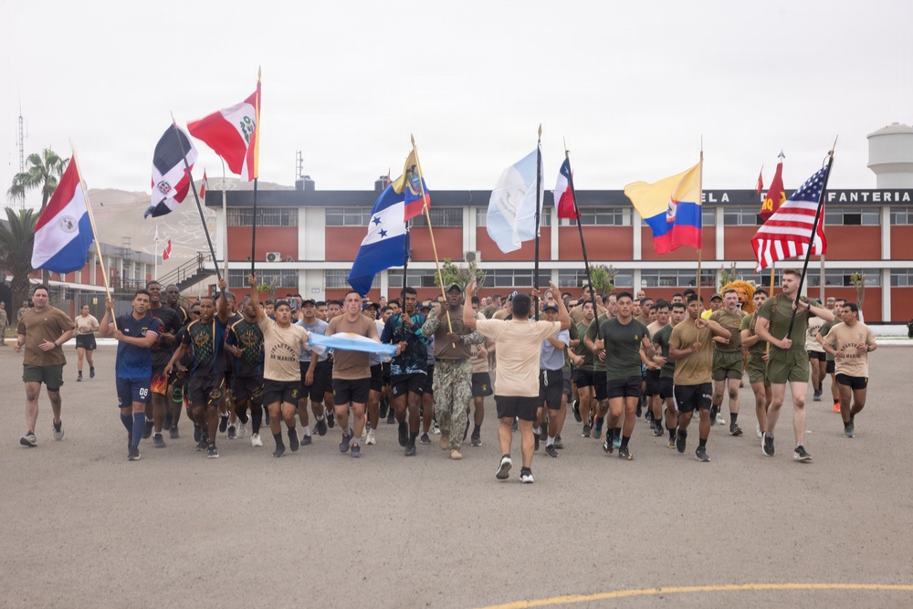 A large group of military personnel from multiple nations are participating in a formation run on a paved parade ground, each carrying their respective national flags. The event appears to be a multinational military exercise or ceremony, likely held at a Latin American military installation based on the building signage reading 'INFANTERIA'. Participants are dressed in various military physical training uniforms representing different countries.
