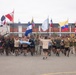 A large group of military personnel from multiple nations are participating in a formation run on a paved parade ground, each carrying their respective national flags. The event appears to be a multinational military exercise or ceremony, likely held at a Latin American military installation based on the building signage reading 'INFANTERIA'. Participants are dressed in various military physical training uniforms representing different countries.