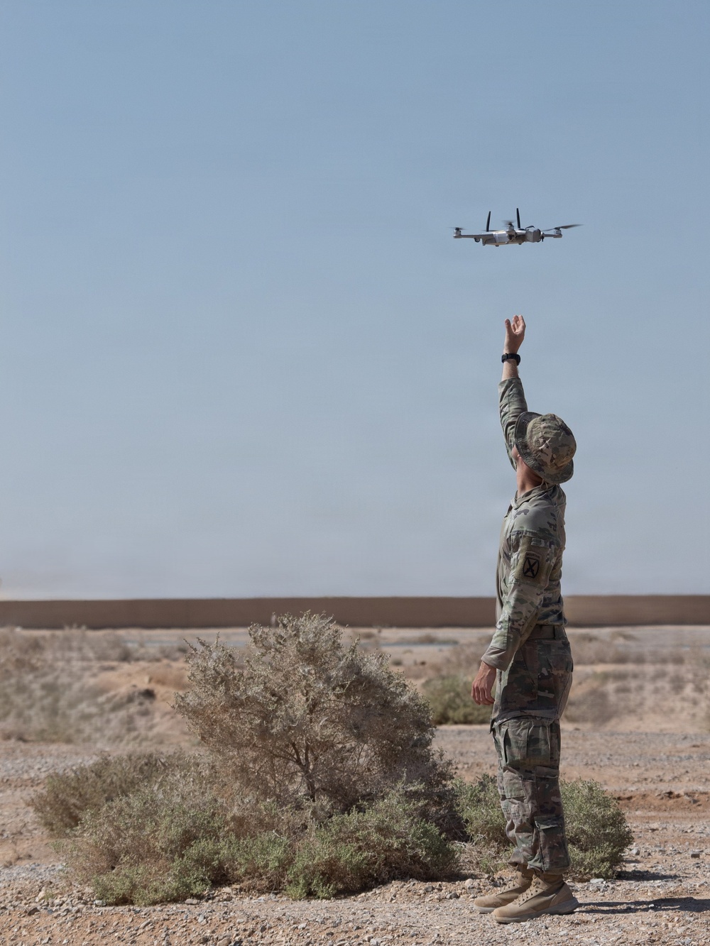 A military service member in OCP (Operational Camouflage Pattern) uniform is launching or catching a small quadrotor/multi-rotor drone in an arid desert environment. The soldier has one arm raised upward toward the drone, which is hovering above. The setting appears to be a remote desert training or operational area with sparse vegetation and sandy terrain.