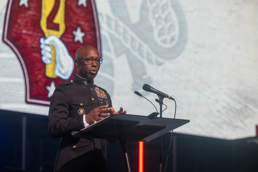 A military officer in dress uniform is speaking at a podium with two microphones during what appears to be a formal ceremony or event. Behind him is a large projection screen displaying what appears to be a military unit insignia featuring a red shield with a yellow torch and stars. The setting suggests an official military function or commemorative event.