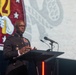 A military officer in dress uniform is speaking at a podium with two microphones during what appears to be a formal ceremony or event. Behind him is a large projection screen displaying what appears to be a military unit insignia featuring a red shield with a yellow torch and stars. The setting suggests an official military function or commemorative event.
