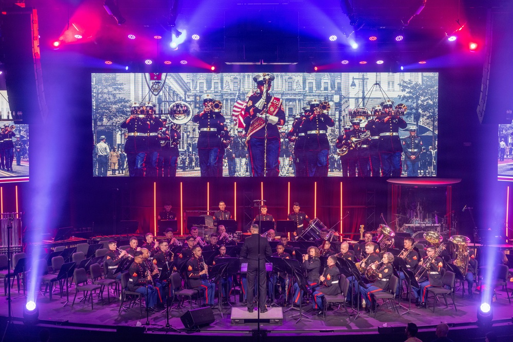 A U.S. Marine Corps band performs on a professionally lit concert stage, with musicians in dress uniforms seated in a semicircular arrangement around a conductor. A large LED screen behind the performers displays a black-and-white historical photograph of Marines marching in a parade. The venue features dramatic red, blue, and purple stage lighting with vertical light columns framing the backdrop.