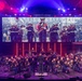 A U.S. Marine Corps band performs on a professionally lit concert stage, with musicians in dress uniforms seated in a semicircular arrangement around a conductor. A large LED screen behind the performers displays a black-and-white historical photograph of Marines marching in a parade. The venue features dramatic red, blue, and purple stage lighting with vertical light columns framing the backdrop.