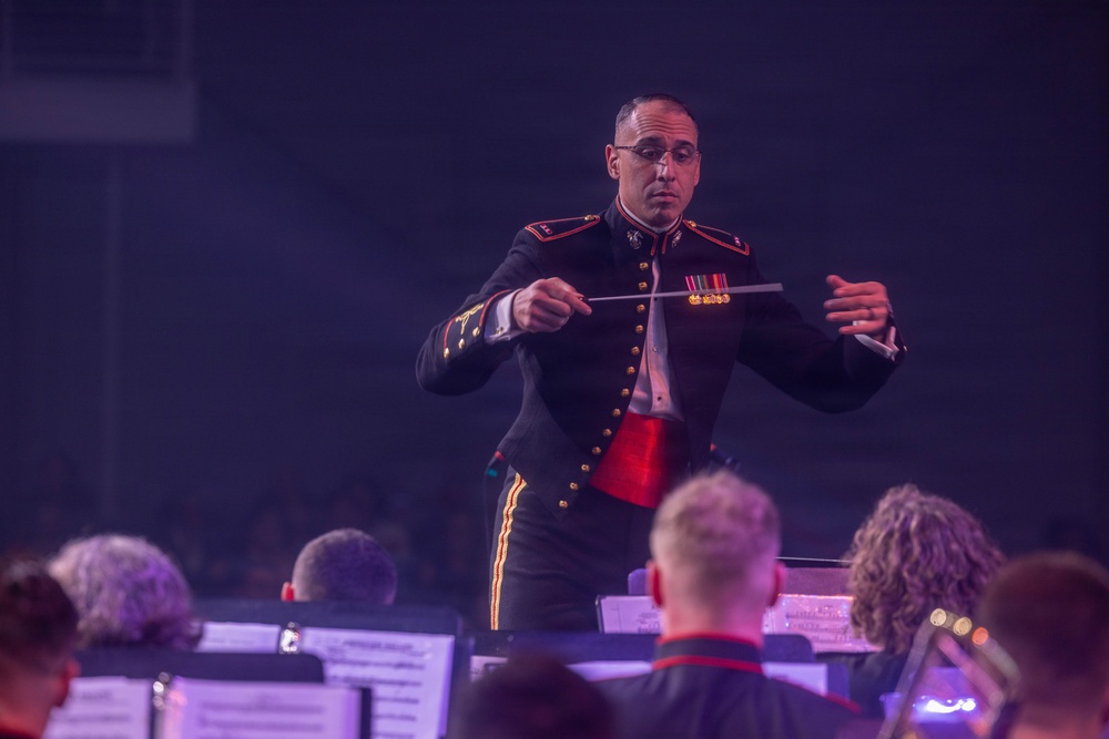 A military band conductor in formal dress uniform is leading a musical ensemble, holding a conductor's baton with both hands extended. He is wearing a dark military uniform with gold buttons, red accents, and service ribbons/medals on his chest. Musicians with sheet music stands are visible in the foreground, slightly out of focus.