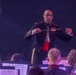 A military band conductor in formal dress uniform is leading a musical ensemble, holding a conductor's baton with both hands extended. He is wearing a dark military uniform with gold buttons, red accents, and service ribbons/medals on his chest. Musicians with sheet music stands are visible in the foreground, slightly out of focus.