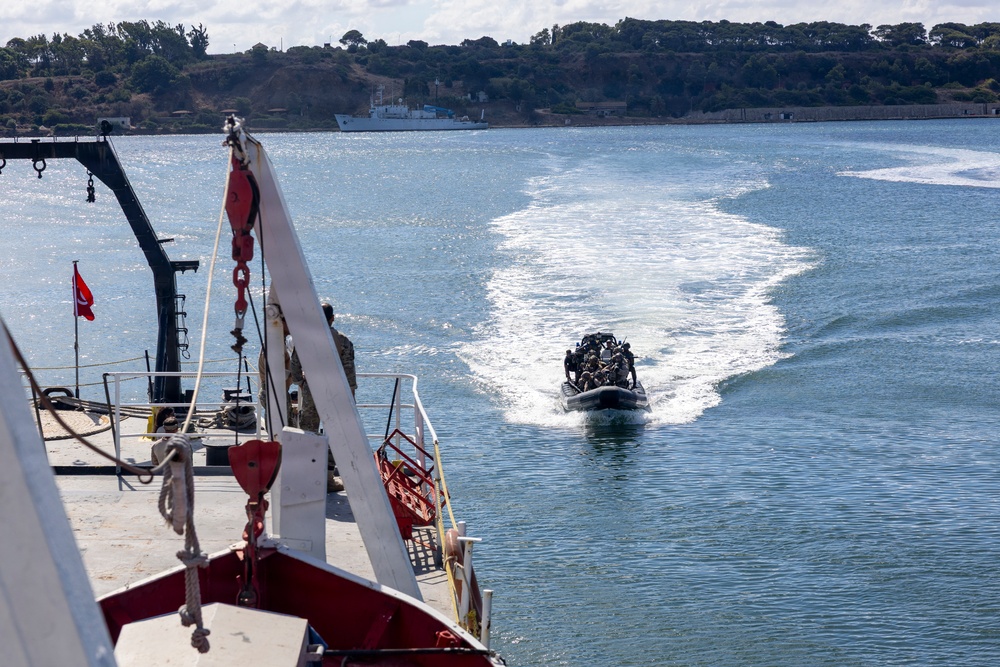 A military or naval operation is underway in a harbor or bay, with a rigid inflatable boat (RIB) carrying armed personnel departing at high speed from a larger vessel. The scene appears to be a tactical exercise or deployment, with the RIB leaving a prominent wake behind it. A naval vessel is visible in the background near a tree-lined shoreline.