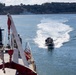 A military or naval operation is underway in a harbor or bay, with a rigid inflatable boat (RIB) carrying armed personnel departing at high speed from a larger vessel. The scene appears to be a tactical exercise or deployment, with the RIB leaving a prominent wake behind it. A naval vessel is visible in the background near a tree-lined shoreline.