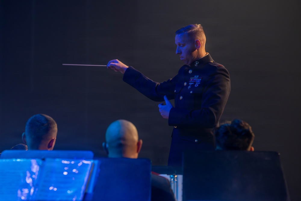 A military conductor in formal dress uniform is leading an orchestra or band, holding a conductor's baton in his right hand. The scene is dramatically lit with blue stage lighting against a dark background. Several musicians are visible from behind in the foreground, seated at music stands.