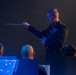 A military conductor in formal dress uniform is leading an orchestra or band, holding a conductor's baton in his right hand. The scene is dramatically lit with blue stage lighting against a dark background. Several musicians are visible from behind in the foreground, seated at music stands.