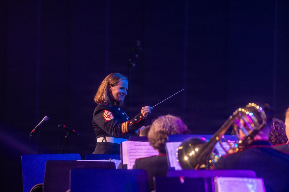 A female military conductor in a dark dress uniform with red chevron insignia is leading an orchestra or band on stage, holding a conductor's baton. She is smiling and appears engaged in the performance. Musicians with brass instruments and sheet music stands are visible in the foreground.