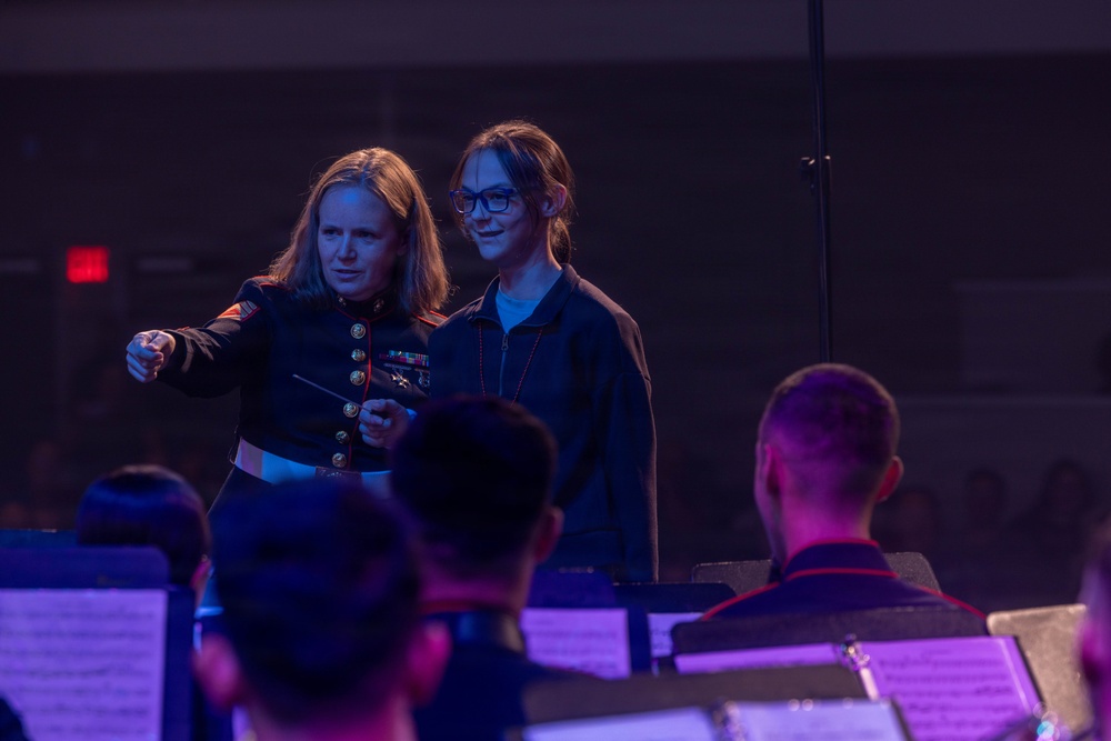 A female U.S. Marine in dress uniform is standing at a conductor's podium, appearing to instruct or guide a young civilian girl in conducting a military band. The Marine holds a conductor's baton and is pointing toward the ensemble, seemingly teaching the youth how to lead the orchestra. Music stands with sheet music are visible in the foreground, with band members seated and facing the conductors.
