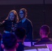 A female U.S. Marine in dress uniform is standing at a conductor's podium, appearing to instruct or guide a young civilian girl in conducting a military band. The Marine holds a conductor's baton and is pointing toward the ensemble, seemingly teaching the youth how to lead the orchestra. Music stands with sheet music are visible in the foreground, with band members seated and facing the conductors.