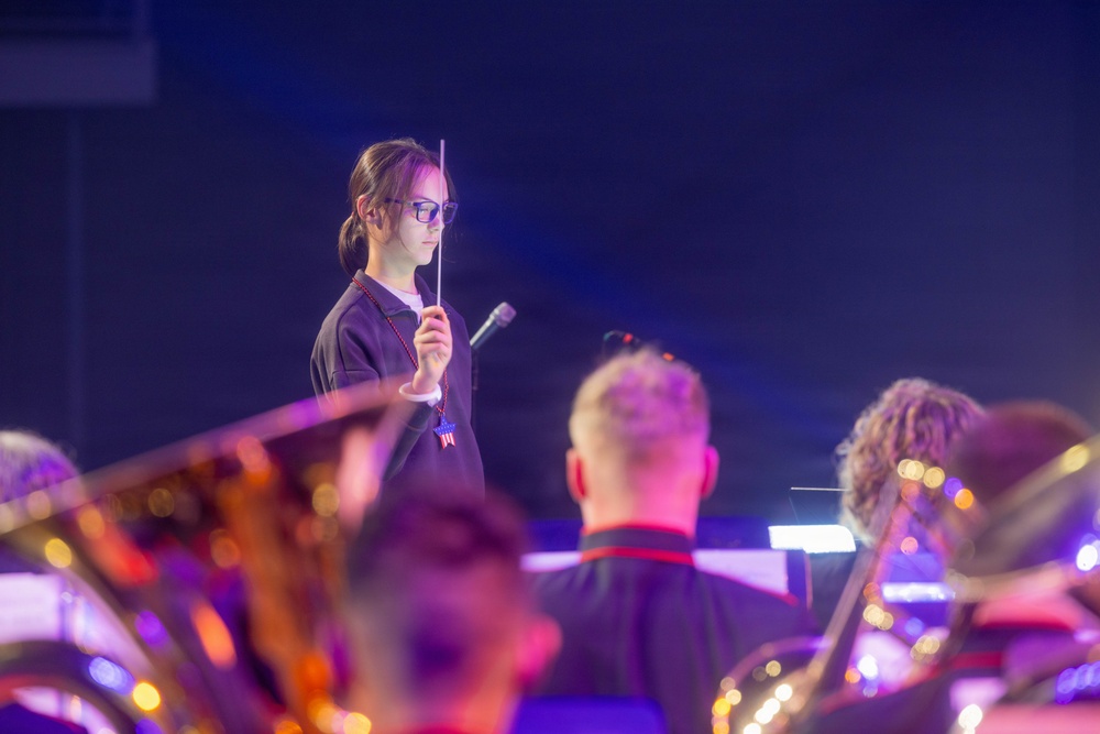 A young female conductor wearing glasses and a dark uniform holds a baton while directing what appears to be a band or orchestra during a performance. She wears a star-shaped pendant and stands elevated above the musicians in the foreground. The scene is lit with dramatic blue and purple stage lighting.