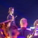A young female conductor wearing glasses and a dark uniform holds a baton while directing what appears to be a band or orchestra during a performance. She wears a star-shaped pendant and stands elevated above the musicians in the foreground. The scene is lit with dramatic blue and purple stage lighting.