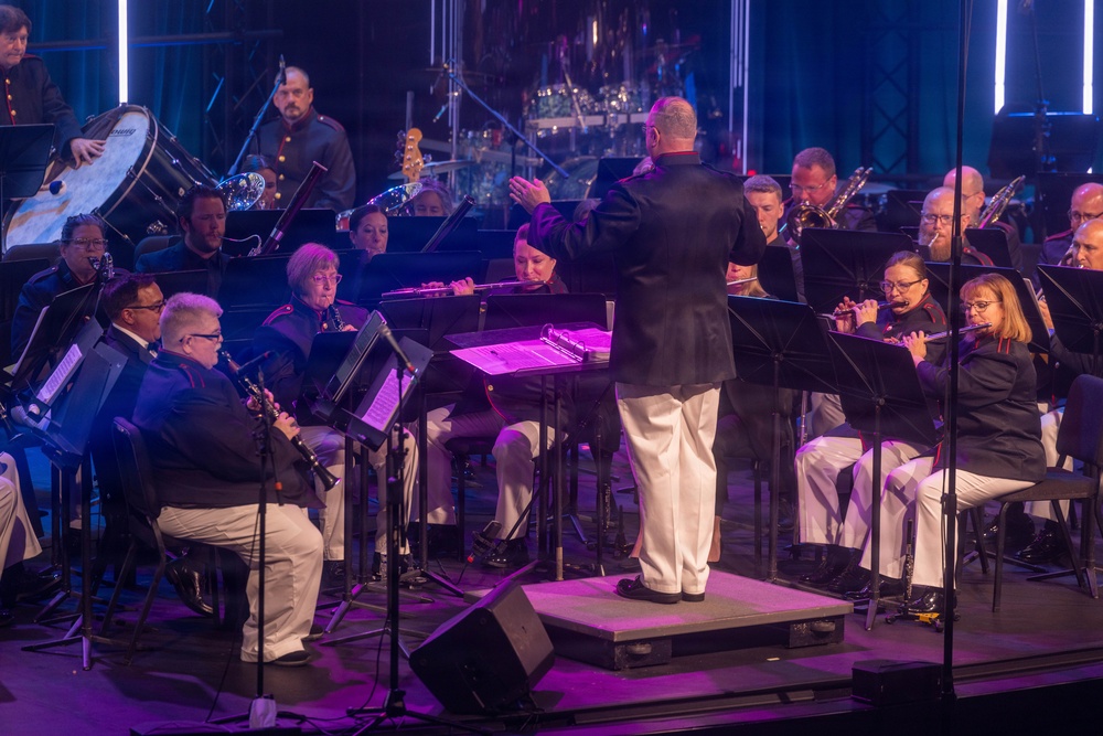 A military band performs on a concert stage under dramatic lighting, led by a conductor standing on a raised podium with his back to the camera. The musicians are dressed in formal military uniforms consisting of dark jackets and white trousers, seated in a semicircular arrangement around the conductor. The performance takes place in a professional venue with purple stage lighting and vertical light columns in the background.