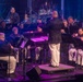 A military band performs on a concert stage under dramatic lighting, led by a conductor standing on a raised podium with his back to the camera. The musicians are dressed in formal military uniforms consisting of dark jackets and white trousers, seated in a semicircular arrangement around the conductor. The performance takes place in a professional venue with purple stage lighting and vertical light columns in the background.