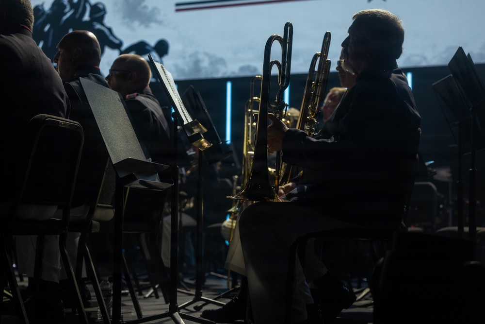 A group of musicians are seated on stage performing in what appears to be an orchestra or concert band setting. The scene is dramatically lit with low, moody lighting, highlighting brass instrument players in the foreground. A large projection screen with silhouetted imagery is visible in the background.