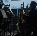 A group of musicians are seated on stage performing in what appears to be an orchestra or concert band setting. The scene is dramatically lit with low, moody lighting, highlighting brass instrument players in the foreground. A large projection screen with silhouetted imagery is visible in the background.