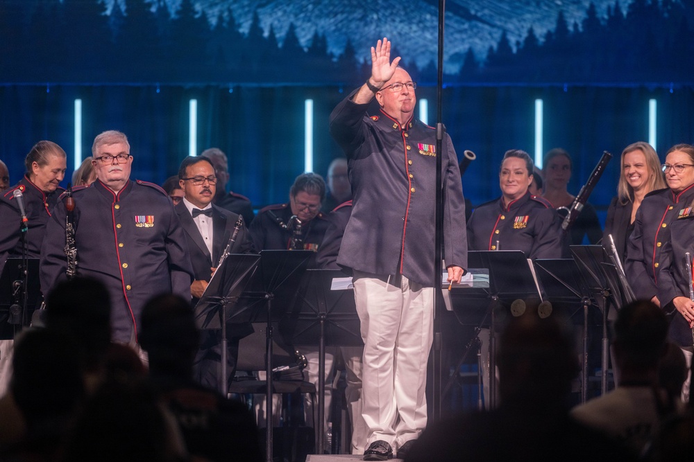 A military band performance on a staged concert venue, featuring musicians in dark navy uniforms with red trim and gold buttons. A central figure, likely the conductor or a featured performer, stands at a microphone with one hand raised, wearing white trousers and a matching uniform jacket adorned with military medals. The backdrop features a blue-toned forest/mountain scenic projection with vertical light columns.