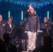 A military band performance on a staged concert venue, featuring musicians in dark navy uniforms with red trim and gold buttons. A central figure, likely the conductor or a featured performer, stands at a microphone with one hand raised, wearing white trousers and a matching uniform jacket adorned with military medals. The backdrop features a blue-toned forest/mountain scenic projection with vertical light columns.
