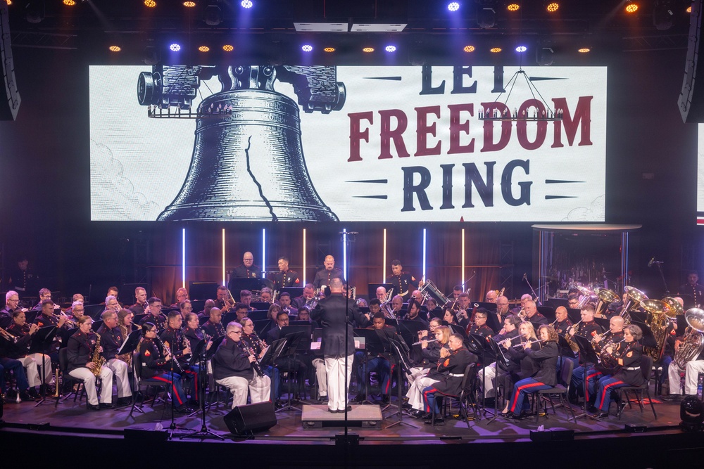 A large military band, appearing to be U.S. Marines based on their distinctive dress uniforms, performs on a well-lit concert stage. A conductor in white trousers leads the ensemble in front of a large projection screen displaying the Liberty Bell and the text 'LET FREEDOM RING'. The performance appears to be a patriotic concert event held in a professional indoor venue.