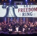 A large military band, appearing to be U.S. Marines based on their distinctive dress uniforms, performs on a well-lit concert stage. A conductor in white trousers leads the ensemble in front of a large projection screen displaying the Liberty Bell and the text 'LET FREEDOM RING'. The performance appears to be a patriotic concert event held in a professional indoor venue.
