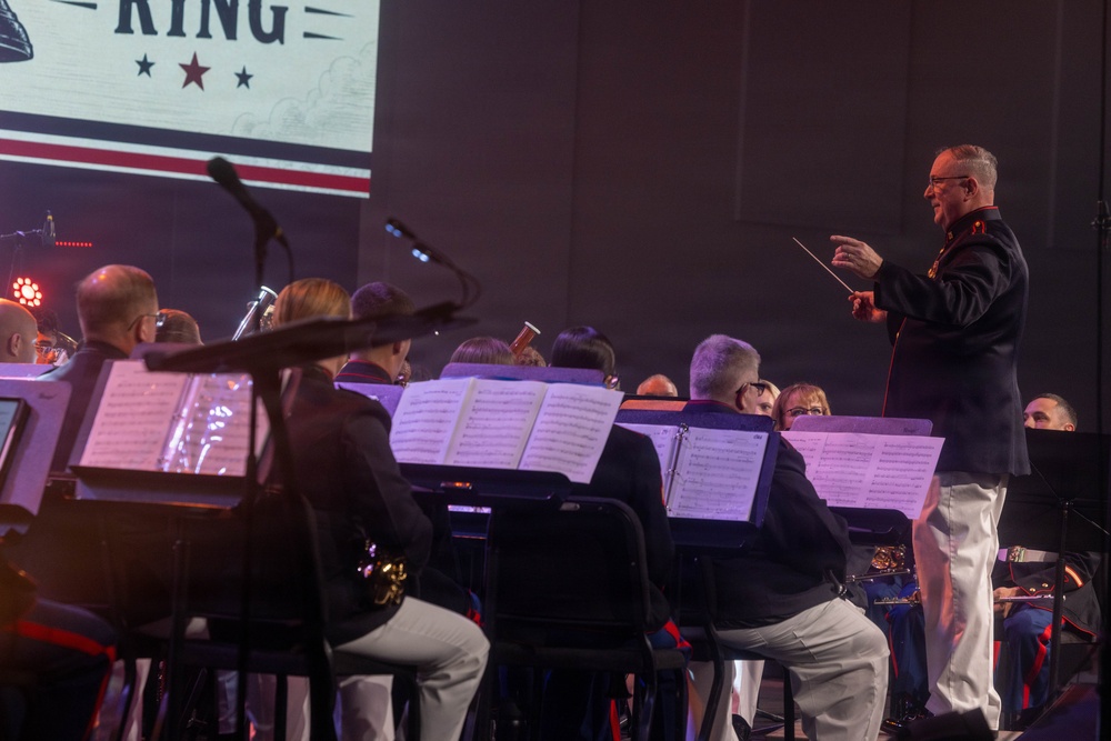A military band performance is taking place on a stage, with a conductor in dress uniform leading the ensemble. The conductor, wearing a dark military jacket with red trim and white trousers, is holding a baton and facing the seated musicians. A projected screen in the background displays partial text reading 'KING' with stars and red/white/blue design elements.