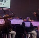 A military band performance is taking place on a stage, with a conductor in dress uniform leading the ensemble. The conductor, wearing a dark military jacket with red trim and white trousers, is holding a baton and facing the seated musicians. A projected screen in the background displays partial text reading 'KING' with stars and red/white/blue design elements.